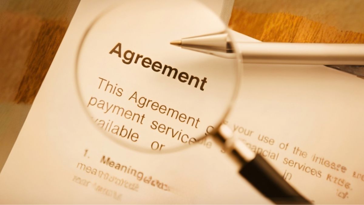 Close-up view of a contract 'Agreement' with a magnifying glass and pen, set in warm, golden lighting on a wooden desk.